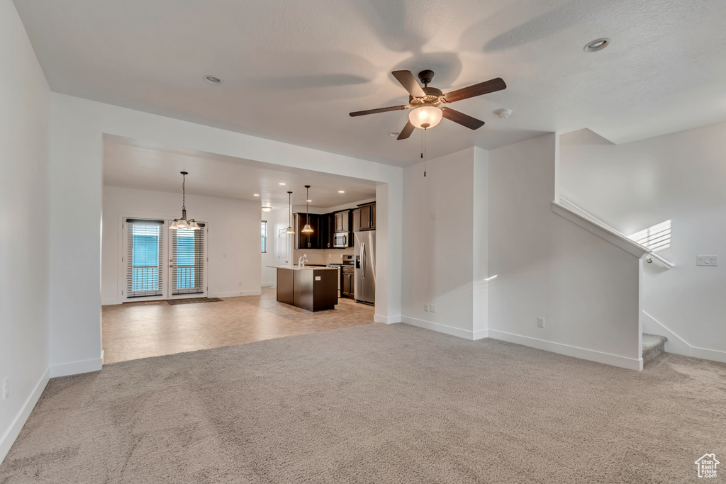 Unfurnished living room with light colored carpet, baseboards, ceiling fan with notable chandelier, and stairs