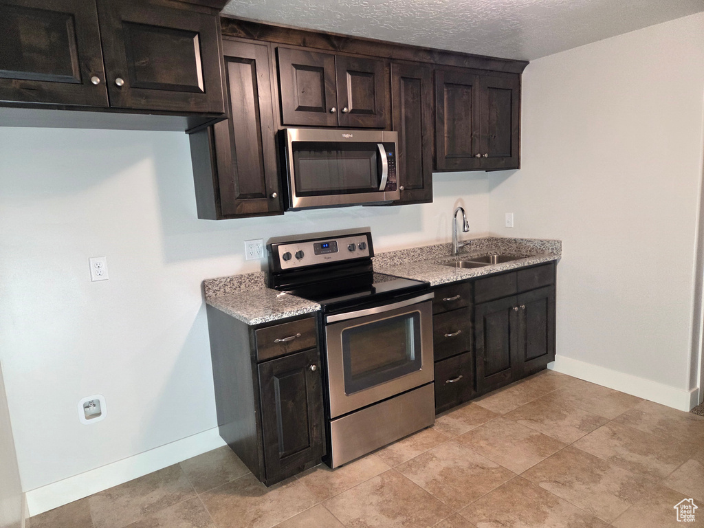 Kitchen with appliances with stainless steel finishes, a sink, baseboards, light stone countertops, and dark brown cabinets