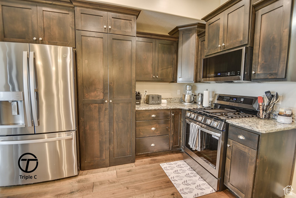 Kitchen with light wood finished floors, dark brown cabinetry, appliances with stainless steel finishes, and light stone countertops