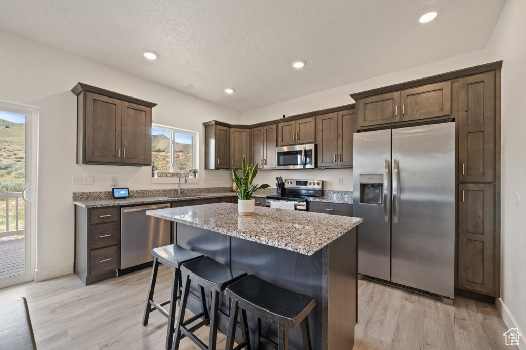 Kitchen with dark brown cabinetry, light wood-type flooring, stainless steel appliances, and light stone countertops