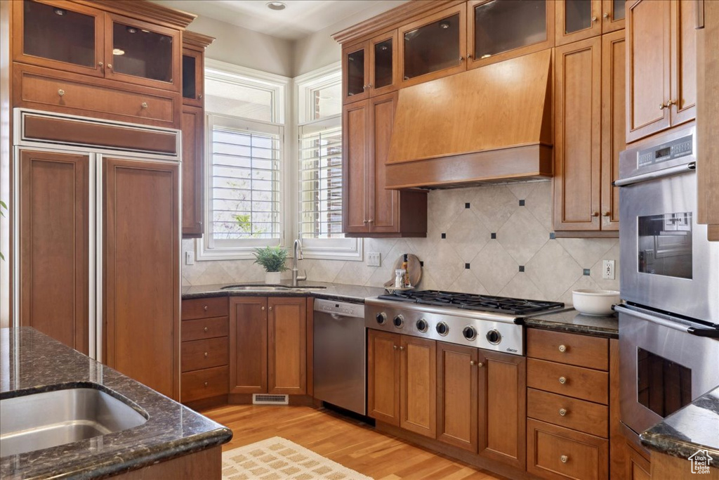 Kitchen with appliances with stainless steel finishes, brown cabinetry, a sink, premium range hood, and light wood-type flooring