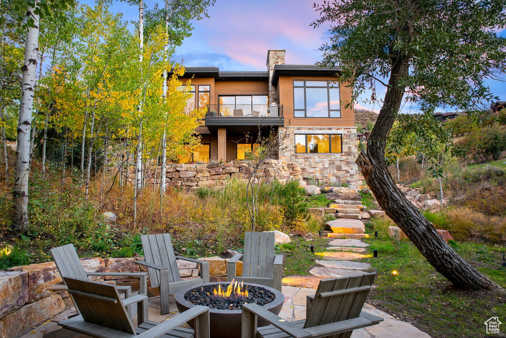 Back of house at dusk featuring stone siding, a balcony, a patio, a chimney, and an outdoor fire pit