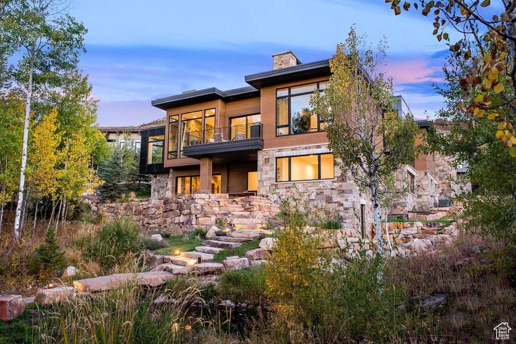 Back of house at dusk featuring stone siding, a chimney, and a balcony