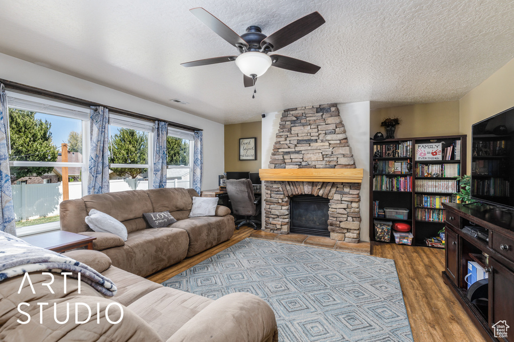 Living room featuring wood finished floors, a textured ceiling, visible vents, ceiling fan, and a stone fireplace