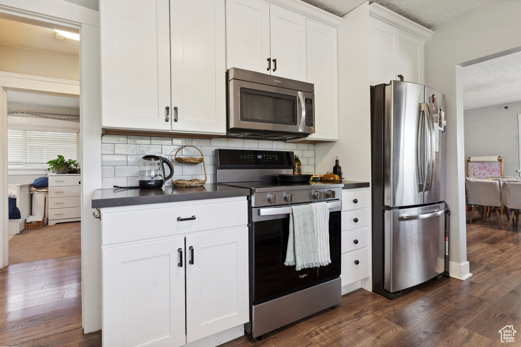 Kitchen with white cabinets, dark wood-type flooring, dark countertops, stainless steel appliances, and tasteful backsplash