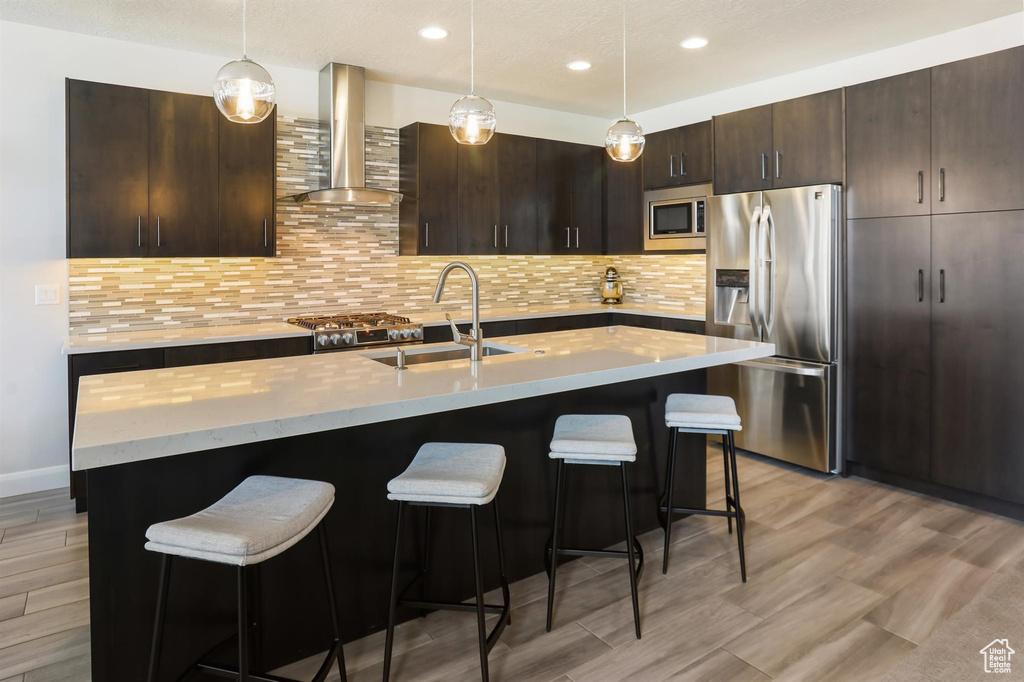Kitchen with dark brown cabinetry, a sink, a kitchen bar, wall chimney range hood, and stainless steel appliances