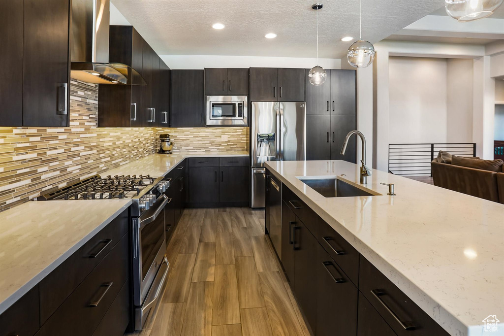 Kitchen with light stone countertops, light wood-style flooring, a sink, wall chimney range hood, and appliances with stainless steel finishes