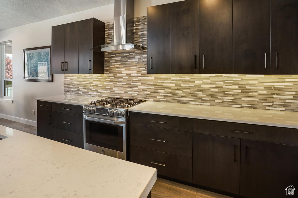 Kitchen with baseboards, stainless steel gas stove, dark brown cabinetry, decorative backsplash, and wall chimney exhaust hood