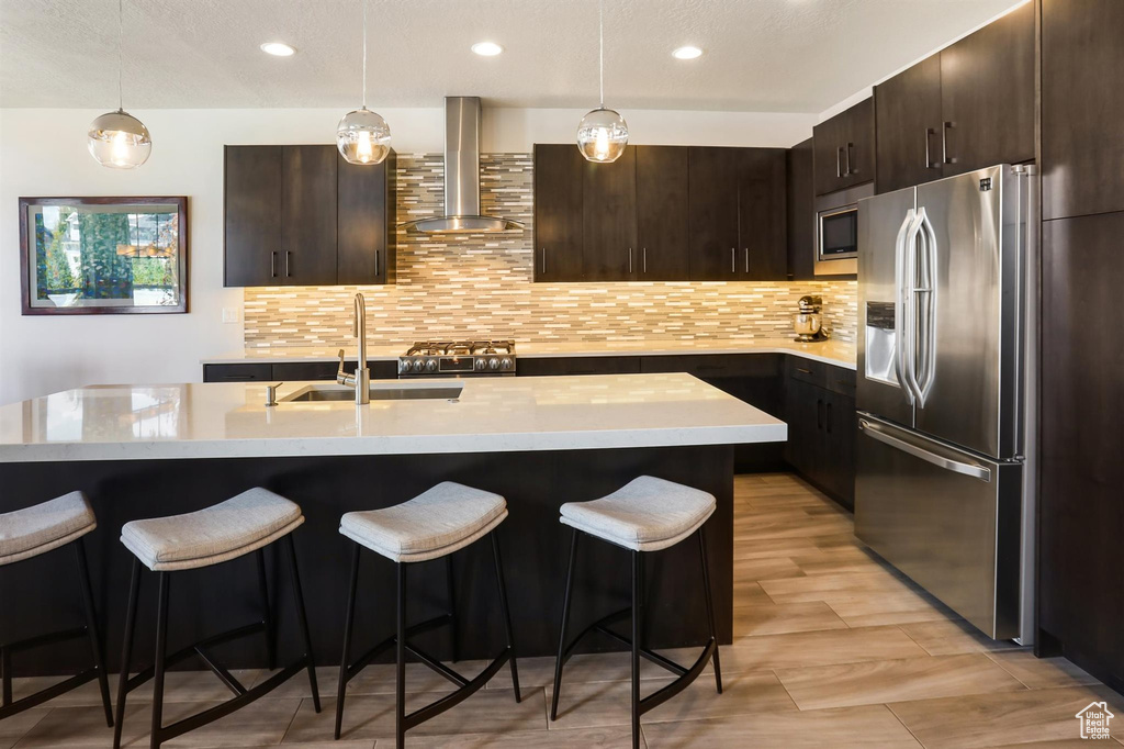 Kitchen featuring a breakfast bar, a sink, wall chimney exhaust hood, stainless steel appliances, and dark brown cabinetry