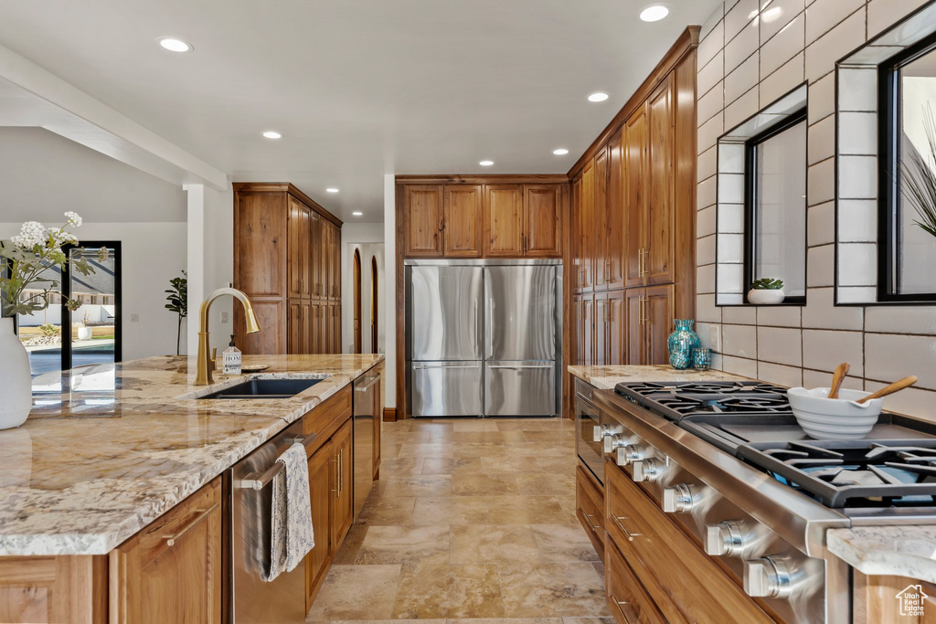 Kitchen with light stone countertops, stainless steel appliances, a sink, backsplash, and brown cabinets