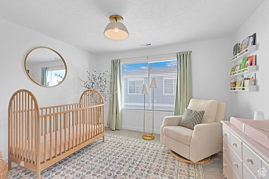 Bedroom with a crib, a textured ceiling, visible vents, baseboards, and light carpet