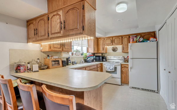 Kitchen with brown cabinetry, white appliances, a peninsula, a kitchen breakfast bar, and light countertops