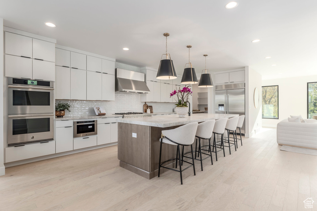 Kitchen featuring a breakfast bar, light wood finished floors, decorative backsplash, wall chimney range hood, and stainless steel appliances
