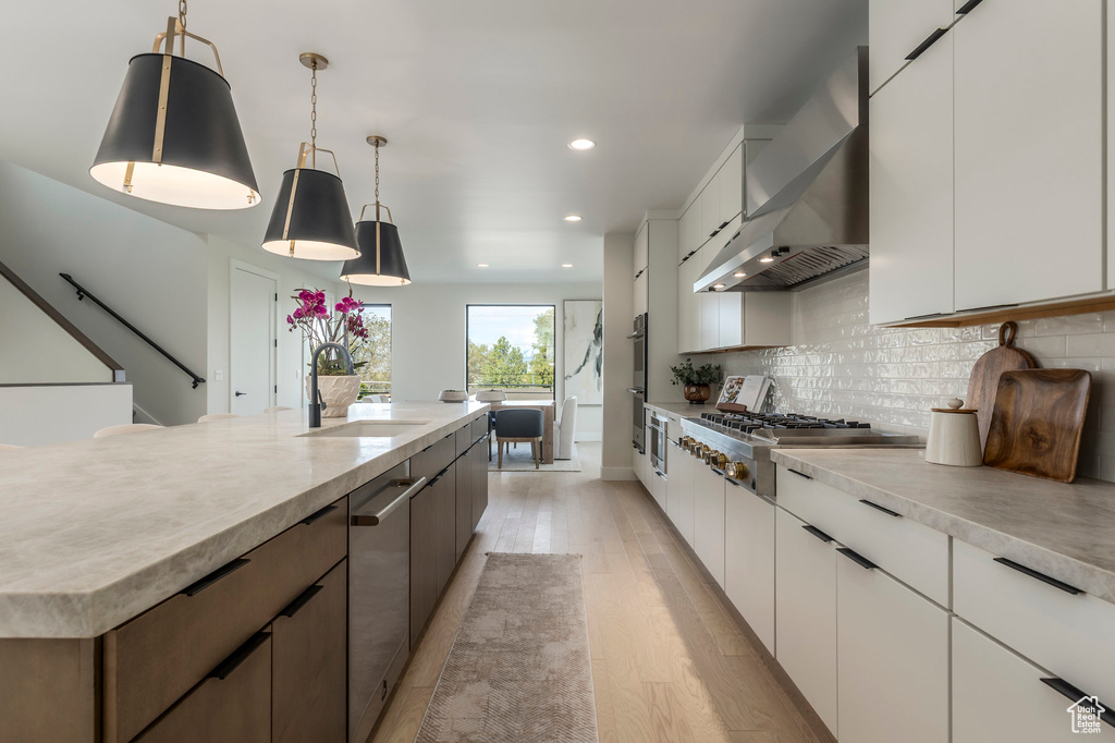 Kitchen featuring a sink, wall chimney exhaust hood, light wood-style floors, light countertops, and appliances with stainless steel finishes