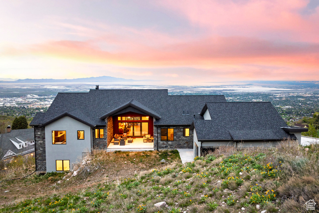 Back of property at dusk featuring stone siding, a mountain view, and a shingled roof