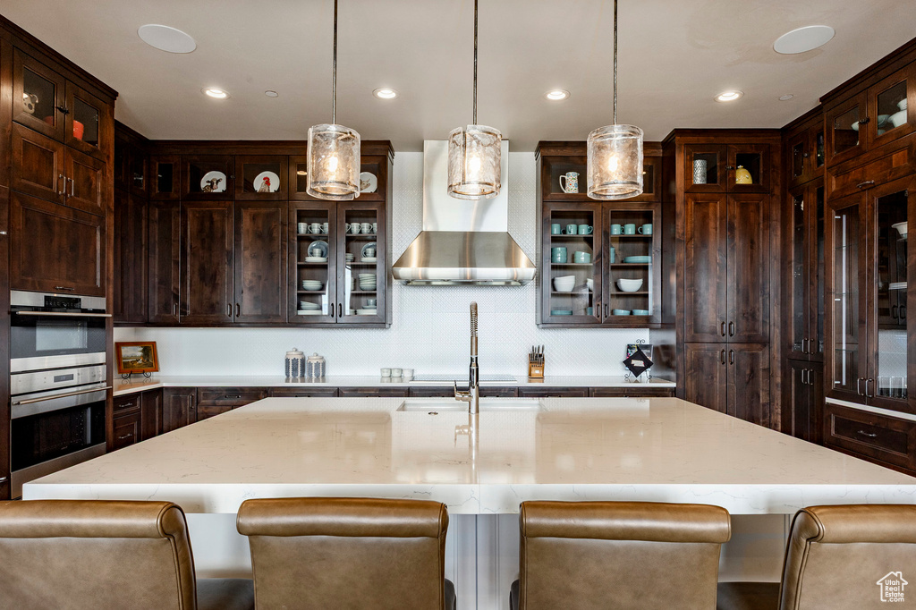 Kitchen with stainless steel double oven, wall chimney exhaust hood, dark brown cabinetry, light stone countertops, and hanging light fixtures