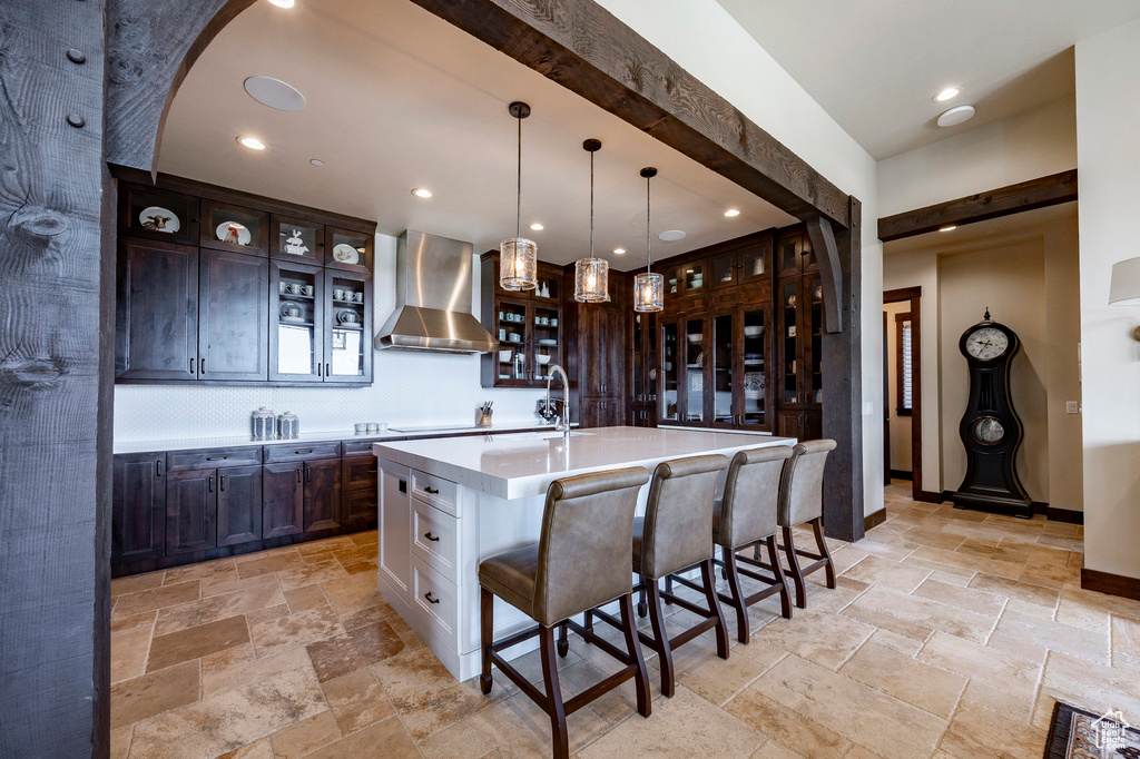 Kitchen with glass insert cabinets, stone tile flooring, a kitchen bar, wall chimney range hood, and light countertops