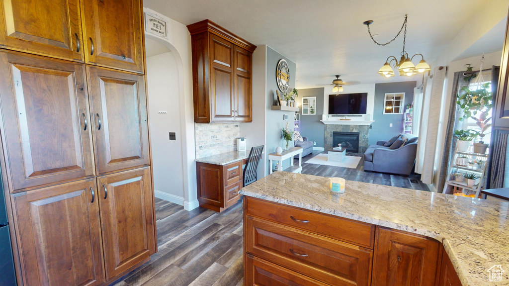 Kitchen featuring light stone countertops, dark wood finished floors, baseboards, ceiling fan with notable chandelier, and a glass covered fireplace