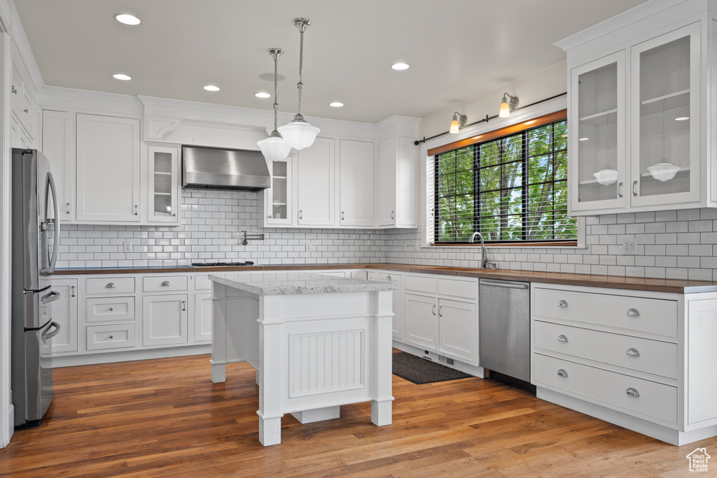 Kitchen with white cabinets, wall chimney range hood, appliances with stainless steel finishes, and backsplash
