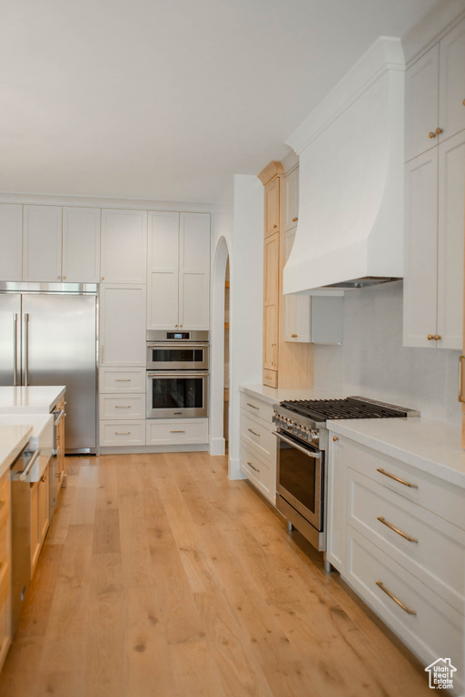 Kitchen featuring high end appliances, arched walkways, light countertops, light wood-style floors, and premium range hood