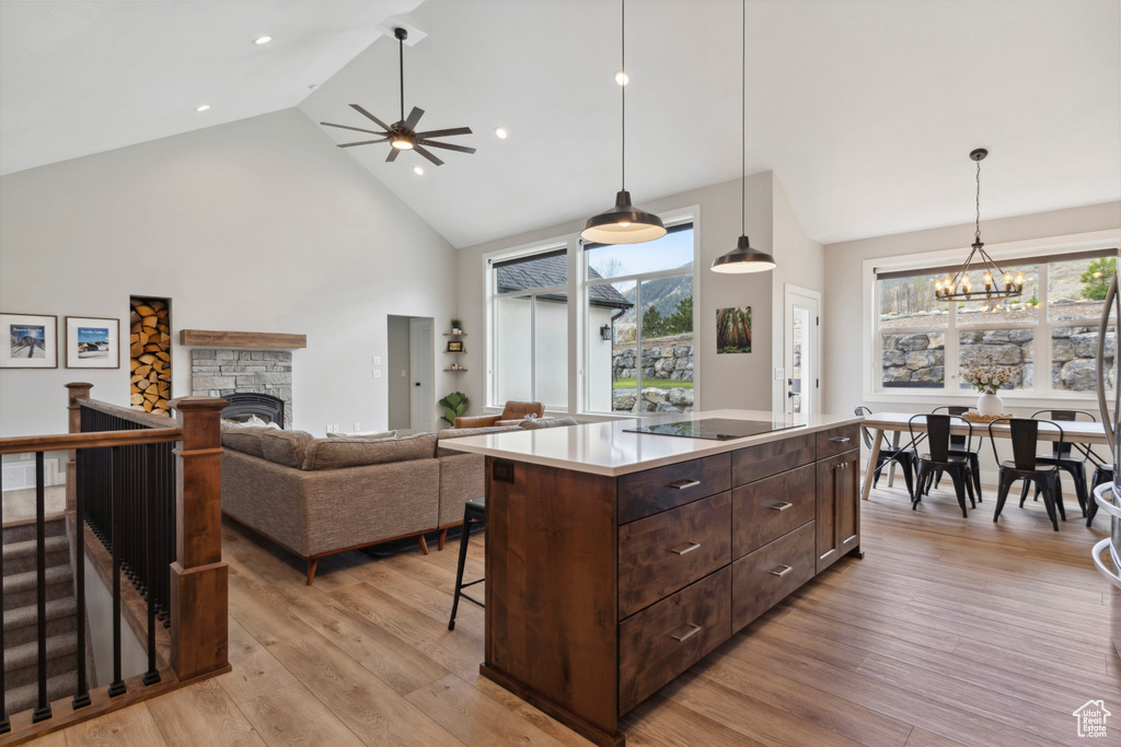 Kitchen with ceiling fan with notable chandelier, high vaulted ceiling, light wood-type flooring, a stone fireplace, and black electric cooktop