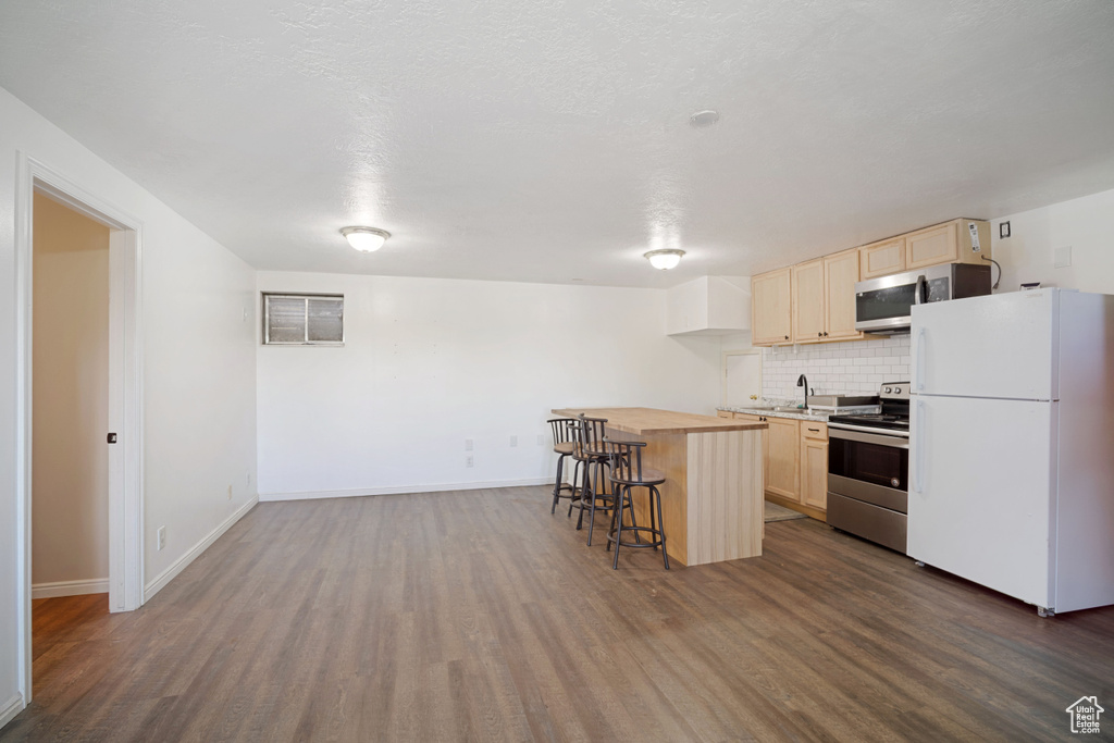 Kitchen featuring a breakfast bar area, light brown cabinetry, appliances with stainless steel finishes, decorative backsplash, and dark wood-style floors