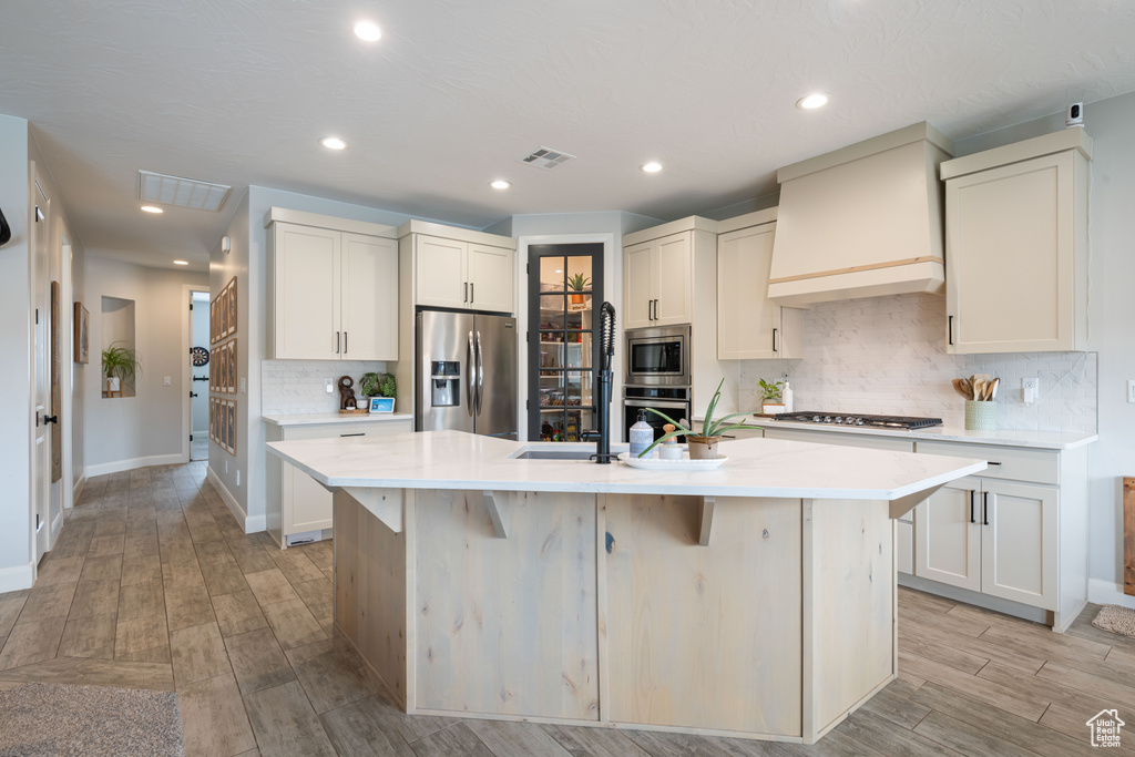 Kitchen featuring appliances with stainless steel finishes, custom range hood, a kitchen island with sink, light countertops, and backsplash