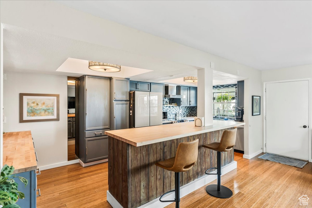 Kitchen with butcher block counters, white fridge, light wood-style flooring, wall chimney range hood, and a kitchen bar