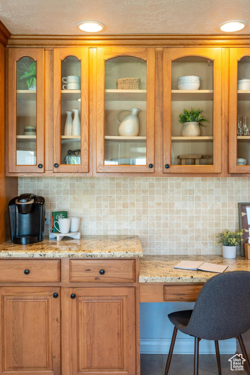 Kitchen featuring glass insert cabinets, backsplash, light stone counters, brown cabinets, and recessed lighting