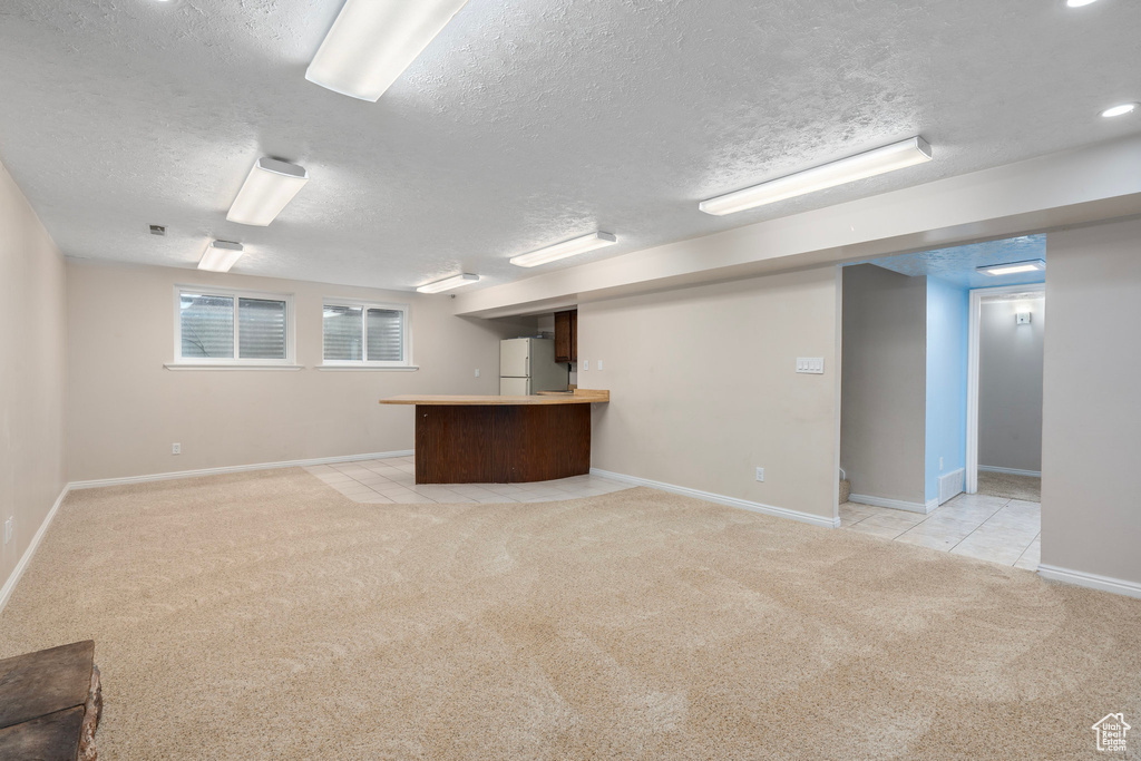 Basement featuring freestanding refrigerator, light colored carpet, a textured ceiling, and baseboards