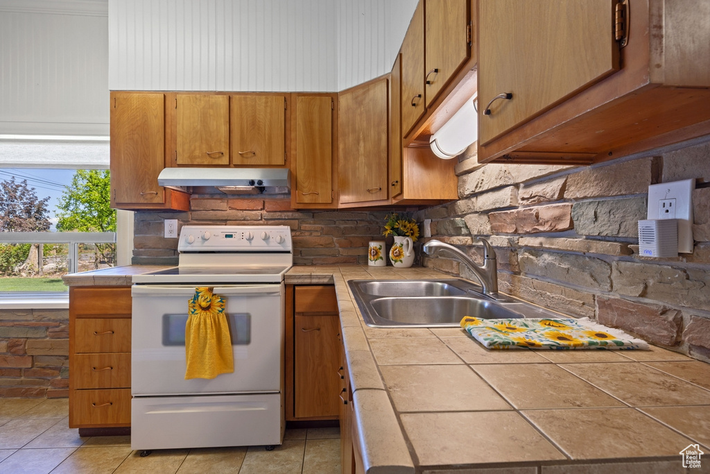 Kitchen featuring electric range, tile countertops, under cabinet range hood, a sink, and brown cabinetry