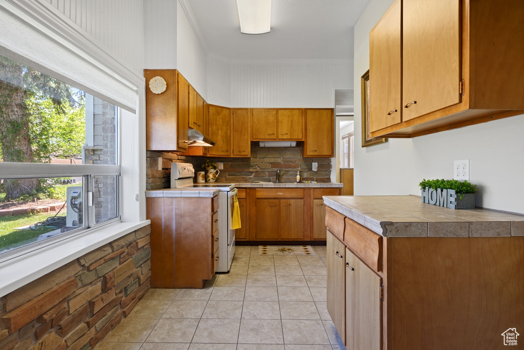 Kitchen with electric range, under cabinet range hood, tasteful backsplash, light tile patterned flooring, and brown cabinetry