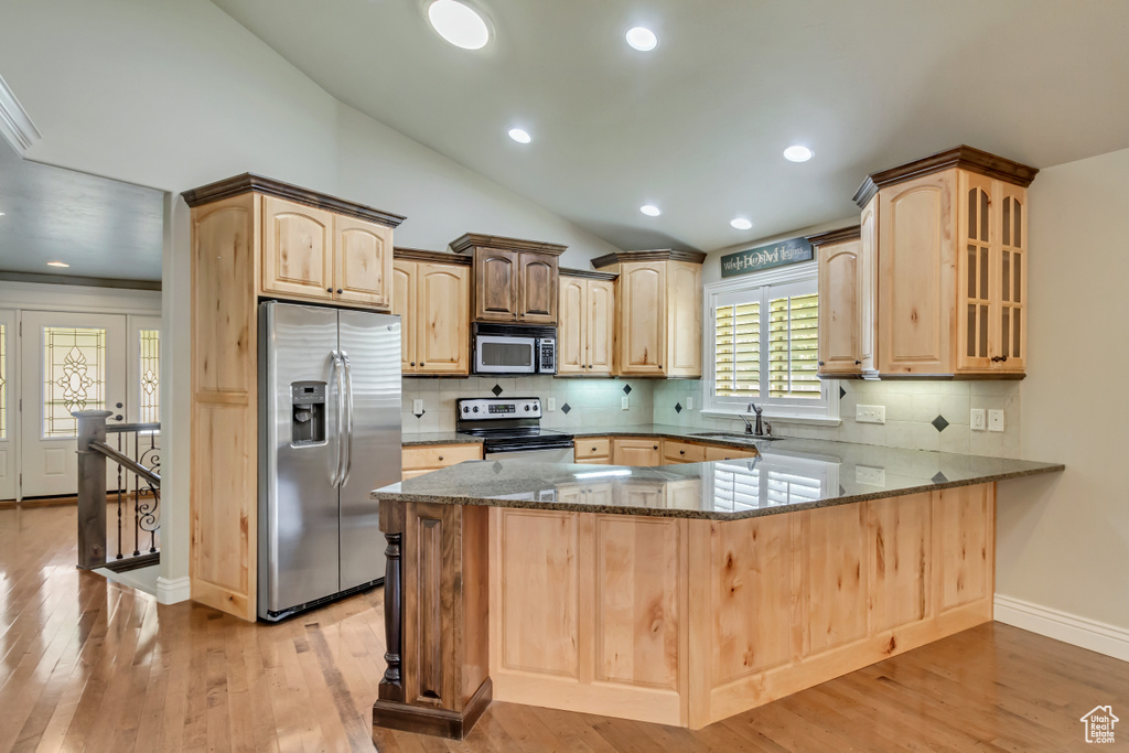 Kitchen featuring appliances with stainless steel finishes, light brown cabinets, dark stone counters, a peninsula, and vaulted ceiling