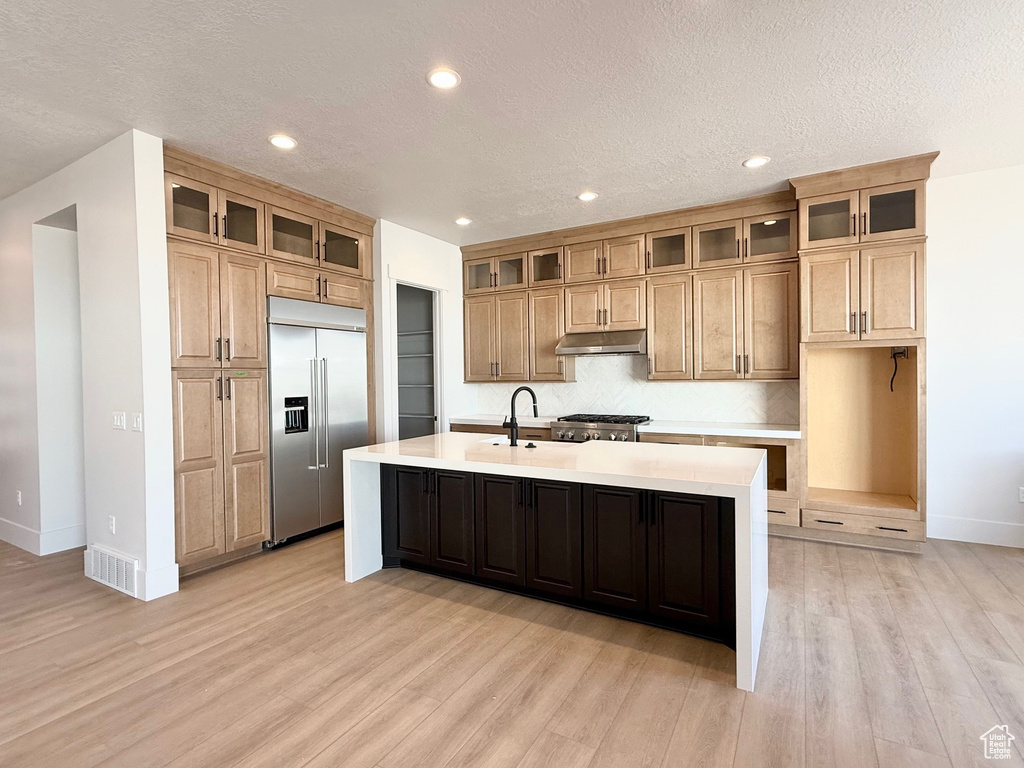 Kitchen featuring built in fridge, a sink, baseboards, a center island with sink, and under cabinet range hood