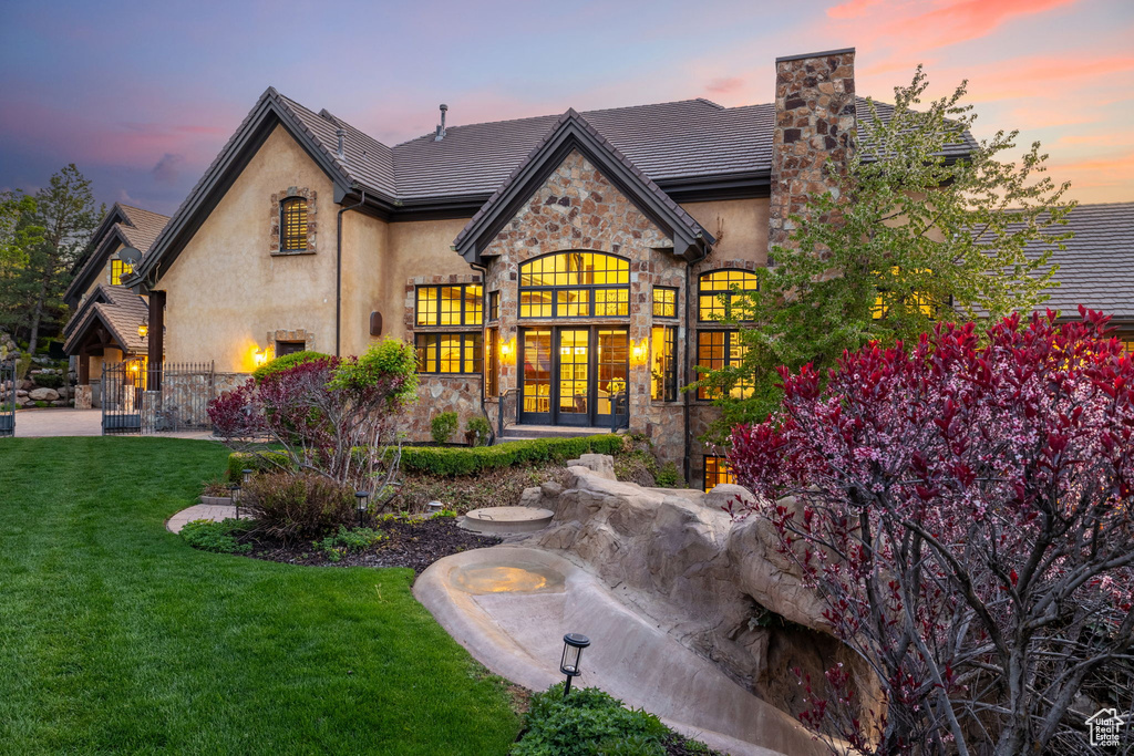 Back of house at dusk featuring a lawn, a chimney, french doors, stucco siding, and stone siding