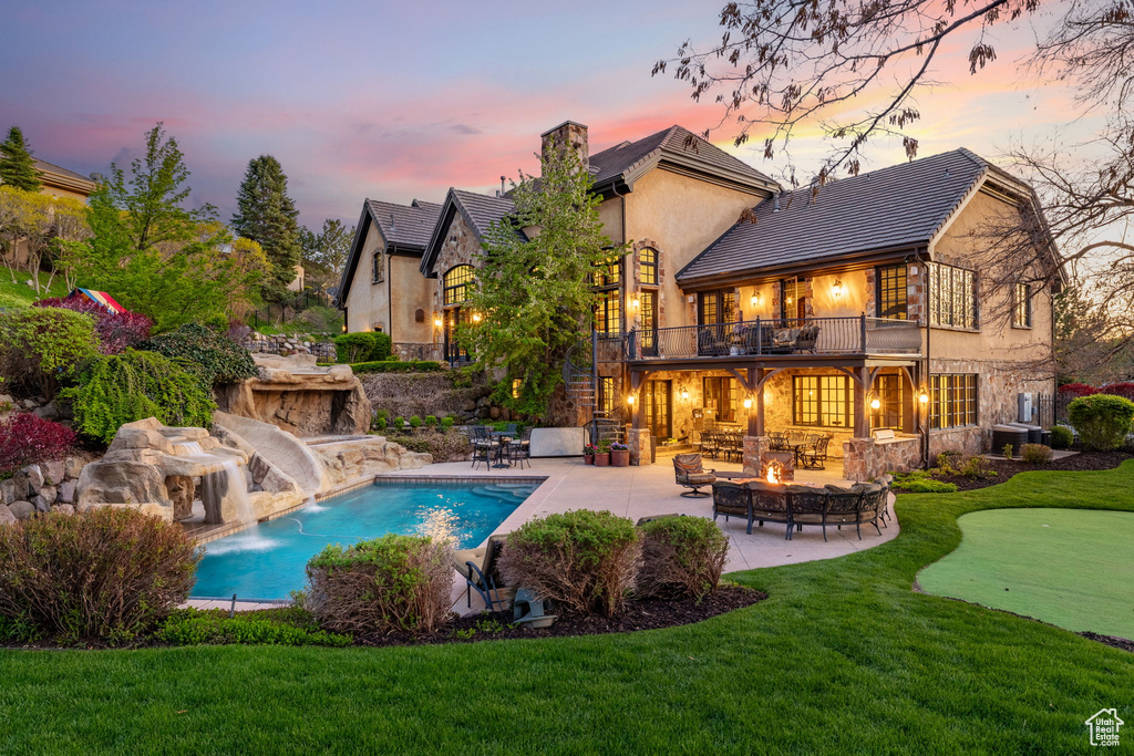 Back of house at dusk featuring a lawn, a patio, stone siding, and a balcony