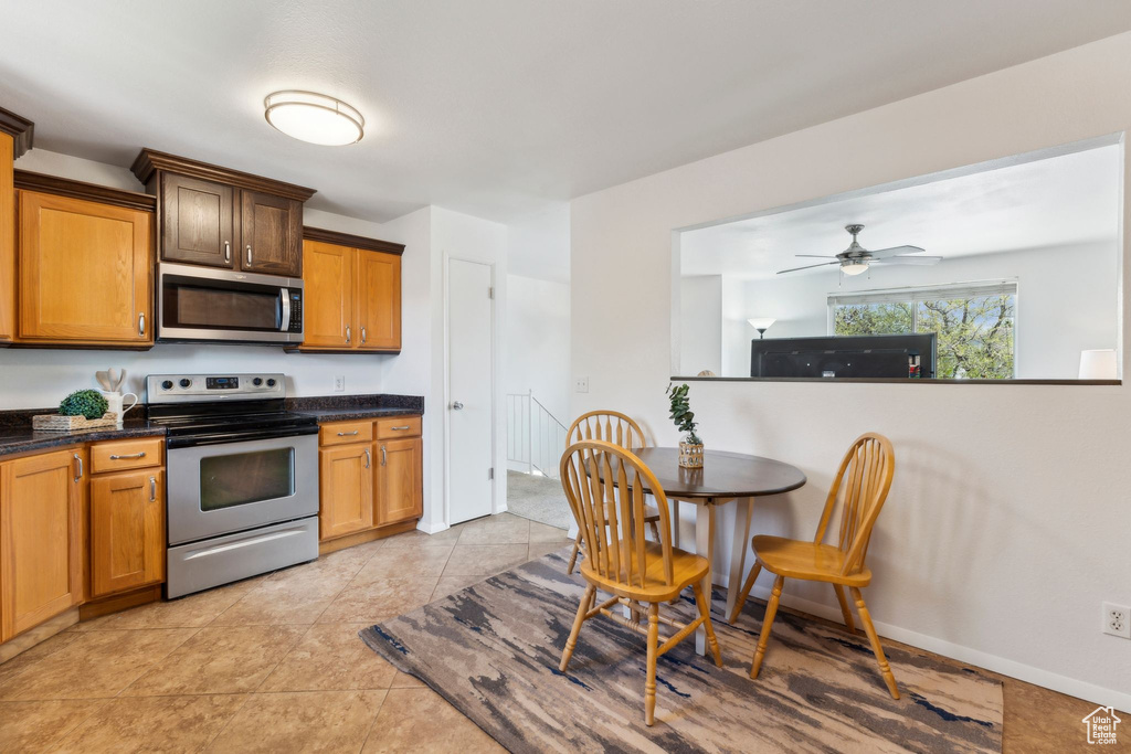 Kitchen with appliances with stainless steel finishes, ceiling fan, light tile patterned flooring, brown cabinets, and dark stone countertops