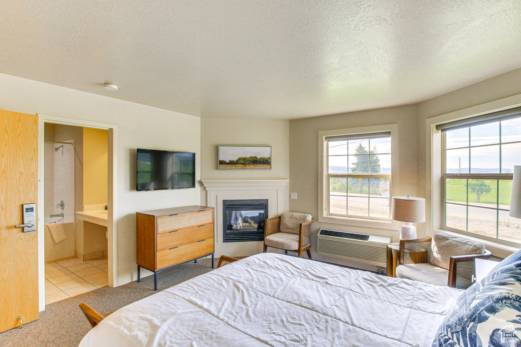 Tiled bedroom featuring ensuite bath, a textured ceiling, a wall unit AC, and a glass covered fireplace