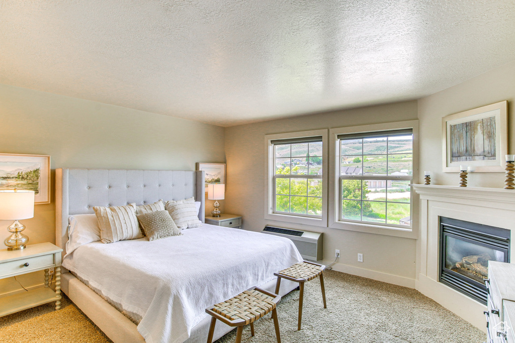 Bedroom featuring light carpet, a textured ceiling, a wall unit AC, and a glass covered fireplace