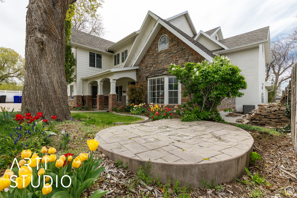 View of front of property with central air condition unit, roof with shingles, a patio area, and stone siding