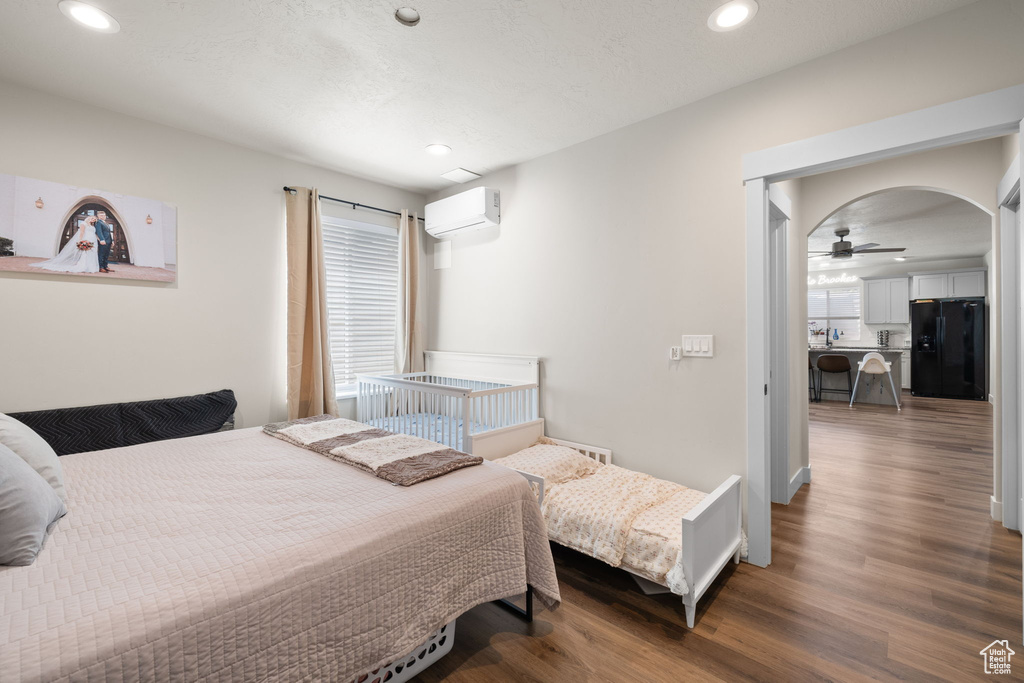 Bedroom featuring an AC wall unit, black refrigerator with ice dispenser, wood finished floors, and multiple windows