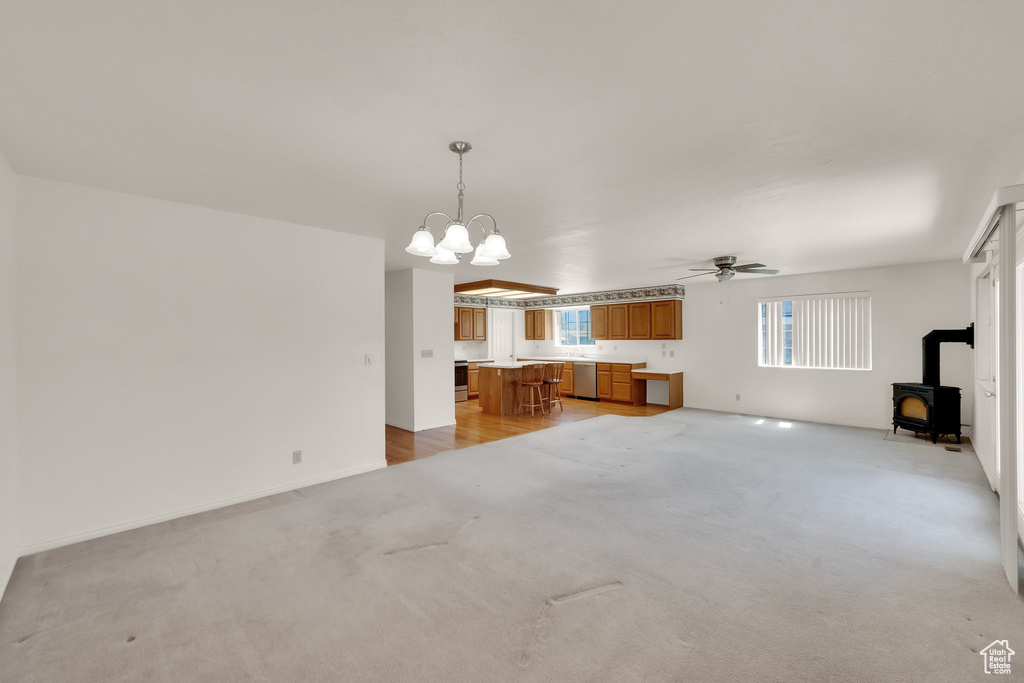 Unfurnished living room featuring a wood stove, ceiling fan, a chandelier, light carpet, and baseboards