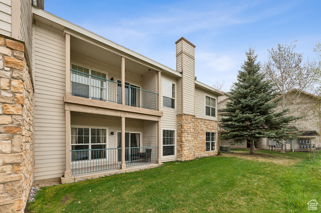 Rear view of house featuring central air condition unit, stone siding, a balcony, a chimney, and a lawn