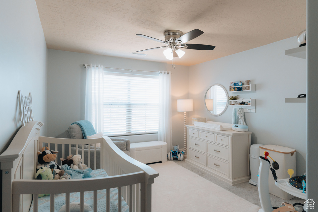 Bedroom featuring light carpet, a ceiling fan, a crib, and a textured ceiling