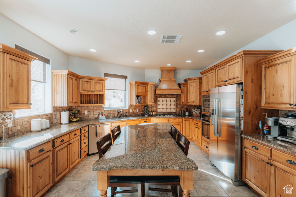 Kitchen featuring stainless steel appliances, premium range hood, decorative backsplash, a center island, and stone countertops
