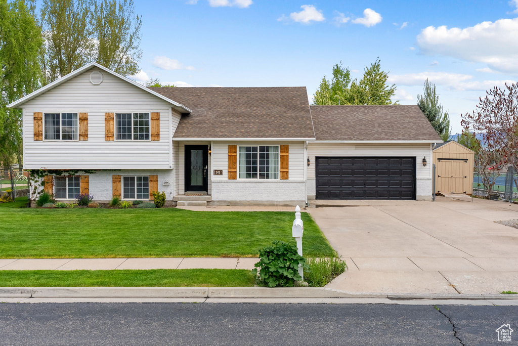 Tri-level home with a garage, a front lawn, driveway, roof with shingles, and a storage unit