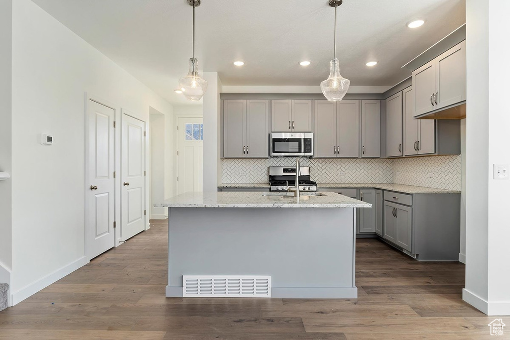 Kitchen featuring gray cabinetry, stainless steel appliances, tasteful backsplash, light stone countertops, and a center island with sink