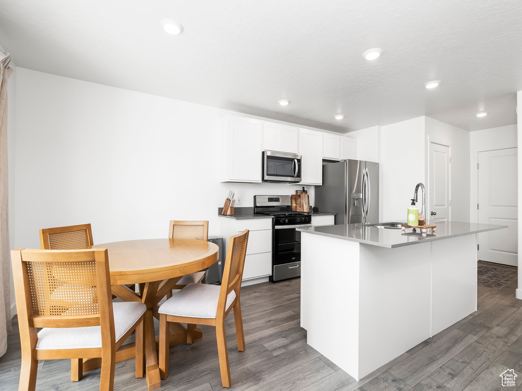 Kitchen with appliances with stainless steel finishes, wood finished floors, a kitchen island with sink, white cabinets, and a sink