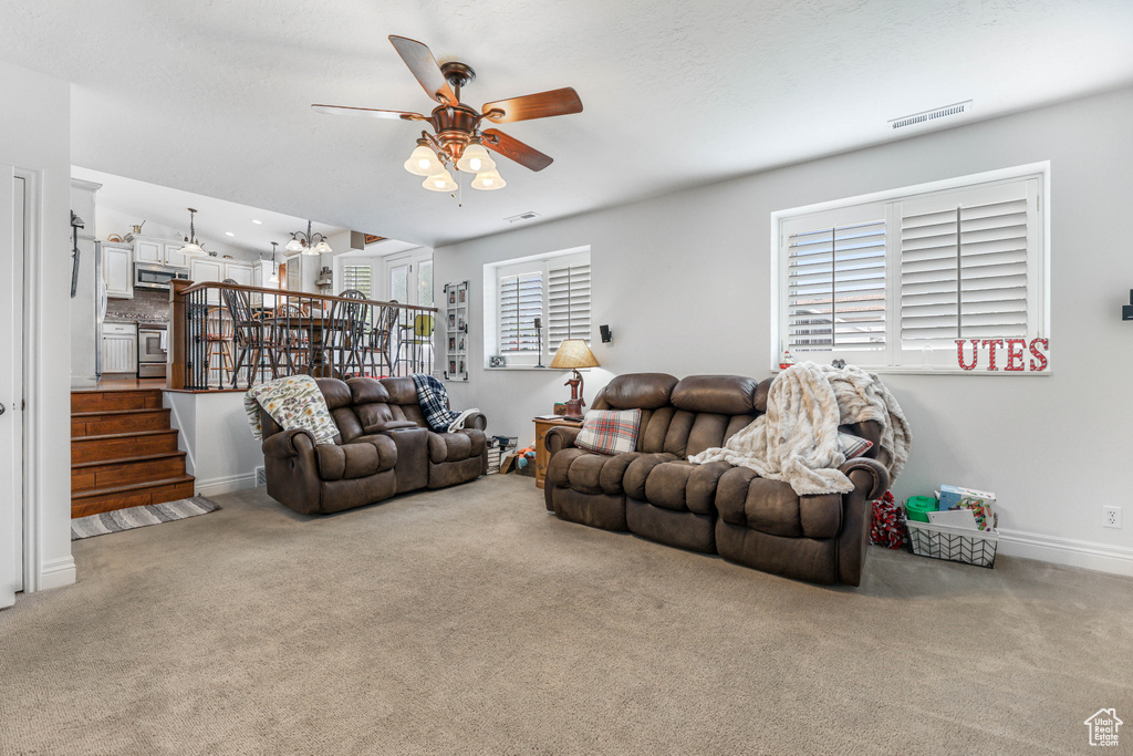 Carpeted living area featuring stairway, ceiling fan with notable chandelier, baseboards, and visible vents