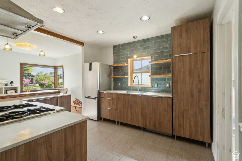 Kitchen with freestanding refrigerator, a sink, wall chimney range hood, decorative backsplash, and brown cabinets